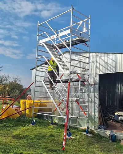Pracownik w żółtym kasku i odblaskowej kamizelce wchodzi biegiem schodowym na aluminiową wieżę jezdną stojącą przy blaszanej hali